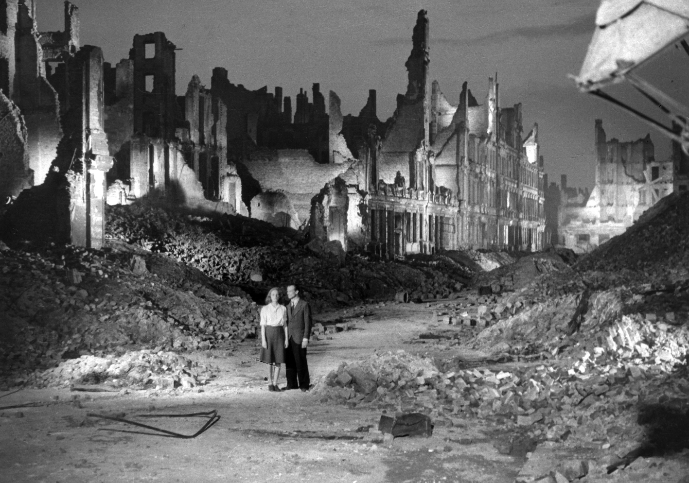 A man and woman are dwarfed by the city rubble surrounding them.